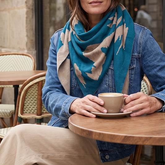 A woman seated at a round wooden table in a cafe, wearing a teal and beige patterned knit scarf, a blue denim jacket, and beige trousers, holding a beige coffee cup and saucer.