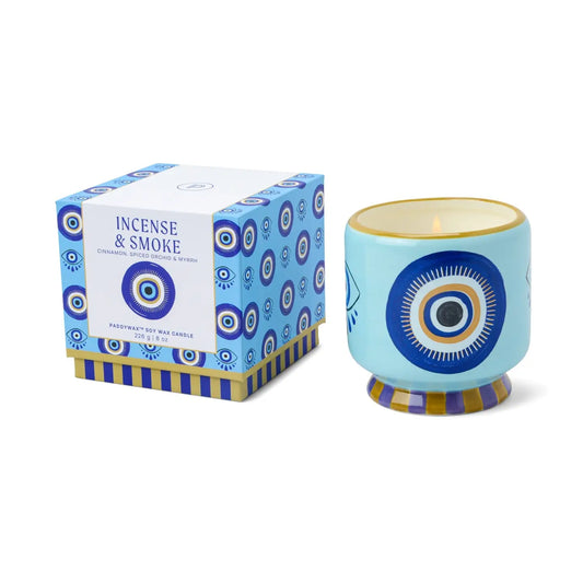 A light blue ceramic candle with a large blue and white evil eye design, shown next to its matching blue and white patterned packaging box on a white background.