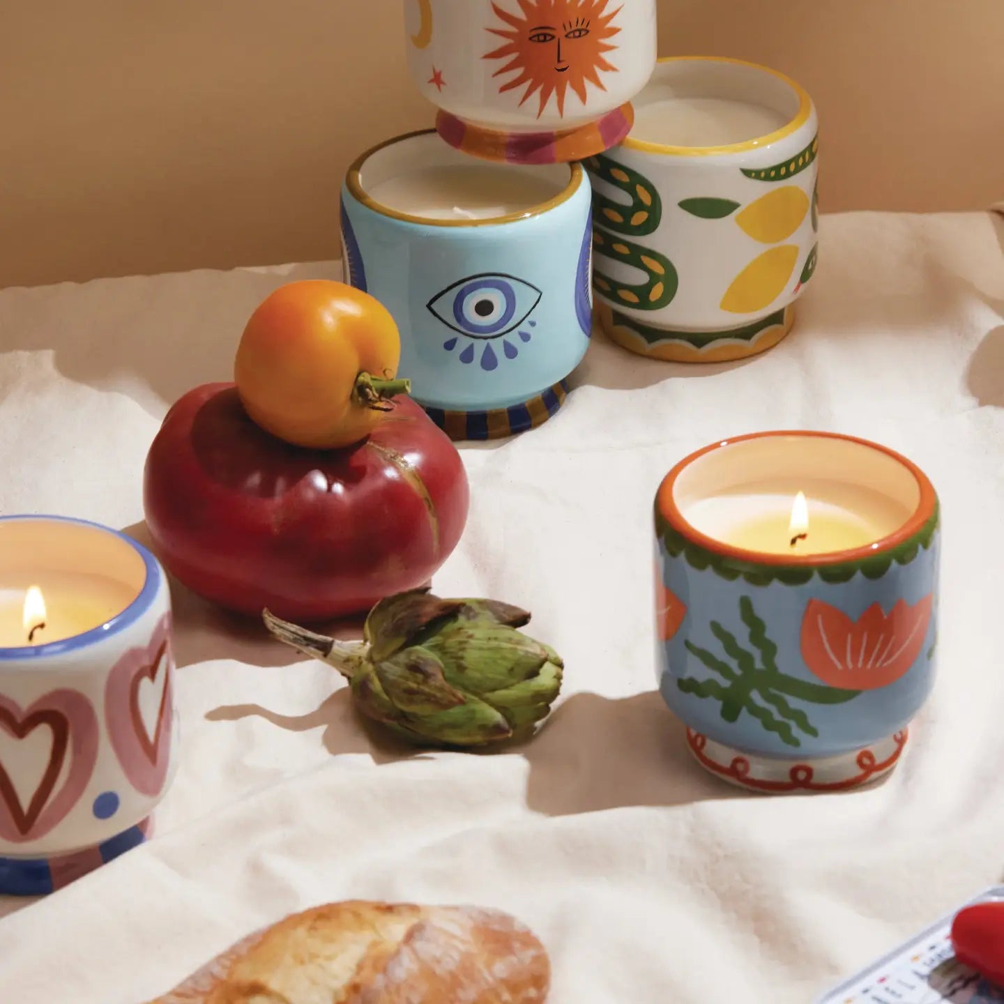 A styled tabletop arrangement of several colorful ceramic candle holders with lit candles, alongside fruit and a loaf of bread on a white cloth. The holders feature hand-painted patterns including hearts, suns, eyes, and botanical motifs in pastel and bright colors.