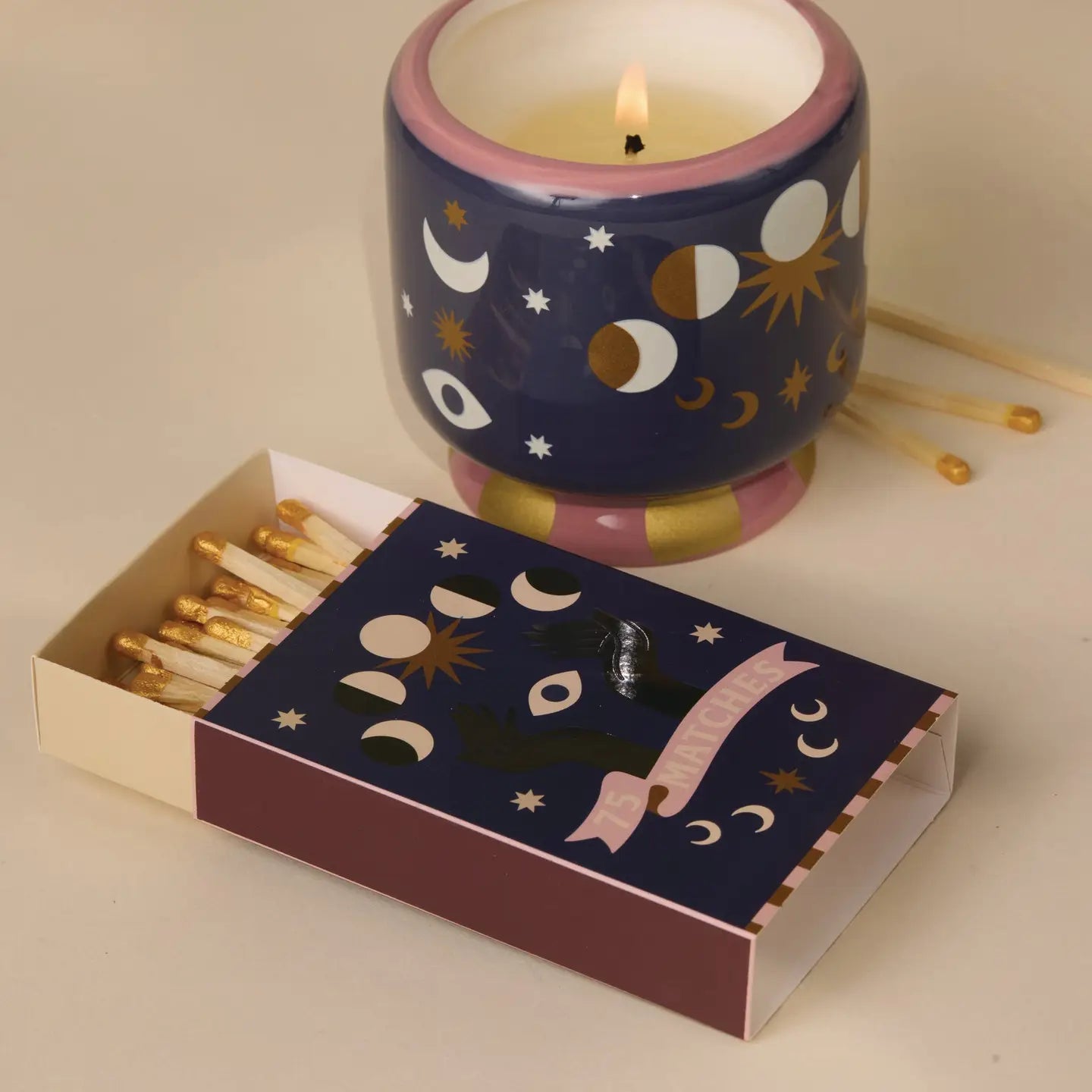 A decorative matchbox and a matching candle holder with a lit candle, both featuring a celestial pattern of moons, stars, and suns on a navy background. The matchbox is open, showing gold-tipped matches, and sits in front of the candle holder on a warm beige surface.