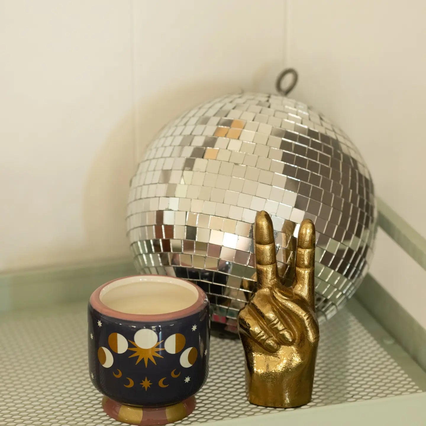 A decorative gold hand sculpture making a peace sign stands next to a small dark blue ceramic cup with moon phase and sun motifs, in front of a large mirrored disco ball on a shelf.