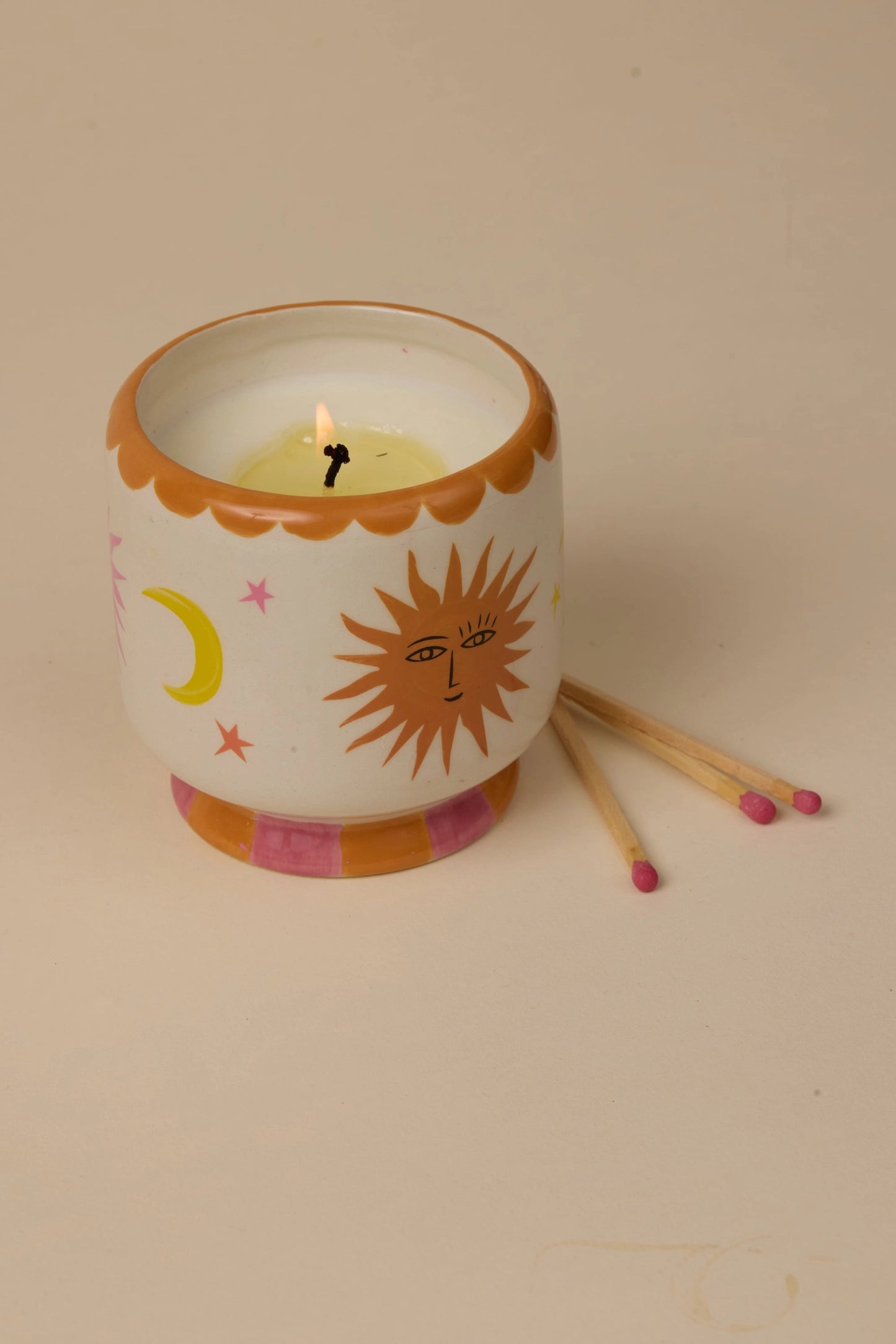 A white ceramic candle holder with a hand-painted sun, moon, and stars design, shown lit with two wooden matches on a beige background.