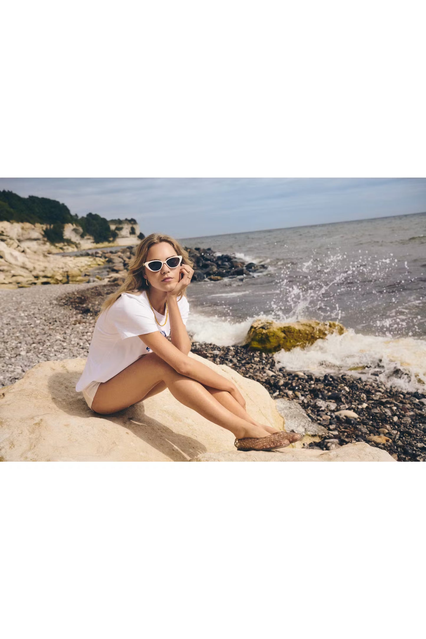 Woman sitting on a rock by the ocean wearing sunglasses.