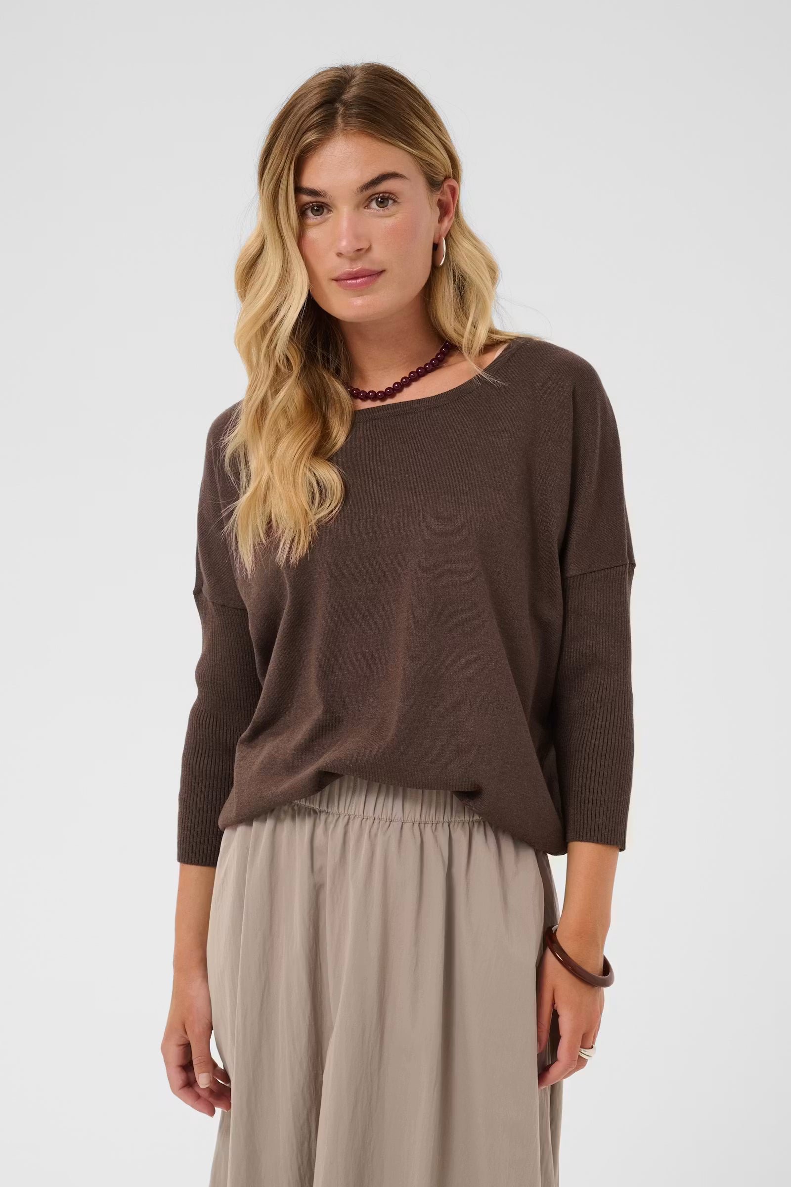 A woman modeling a brown long-sleeve knit top with a wide boat neckline, paired with light beige wide-leg pants, against a plain light gray studio background.