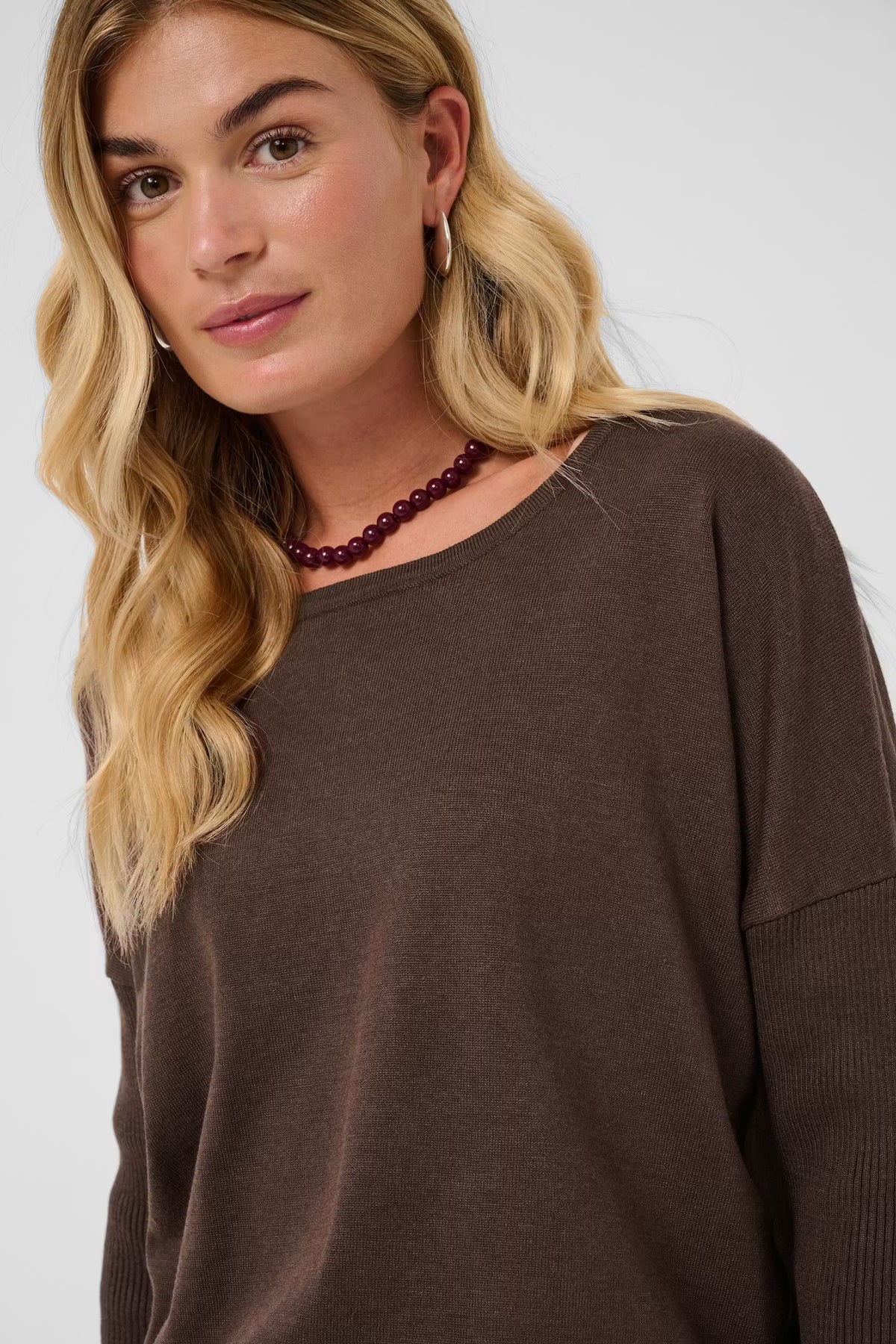 A close-up shot of a woman wearing a brown knit crewneck sweater, featuring her blonde hair, a dark beaded necklace, and the textured sweater against a light gray background.