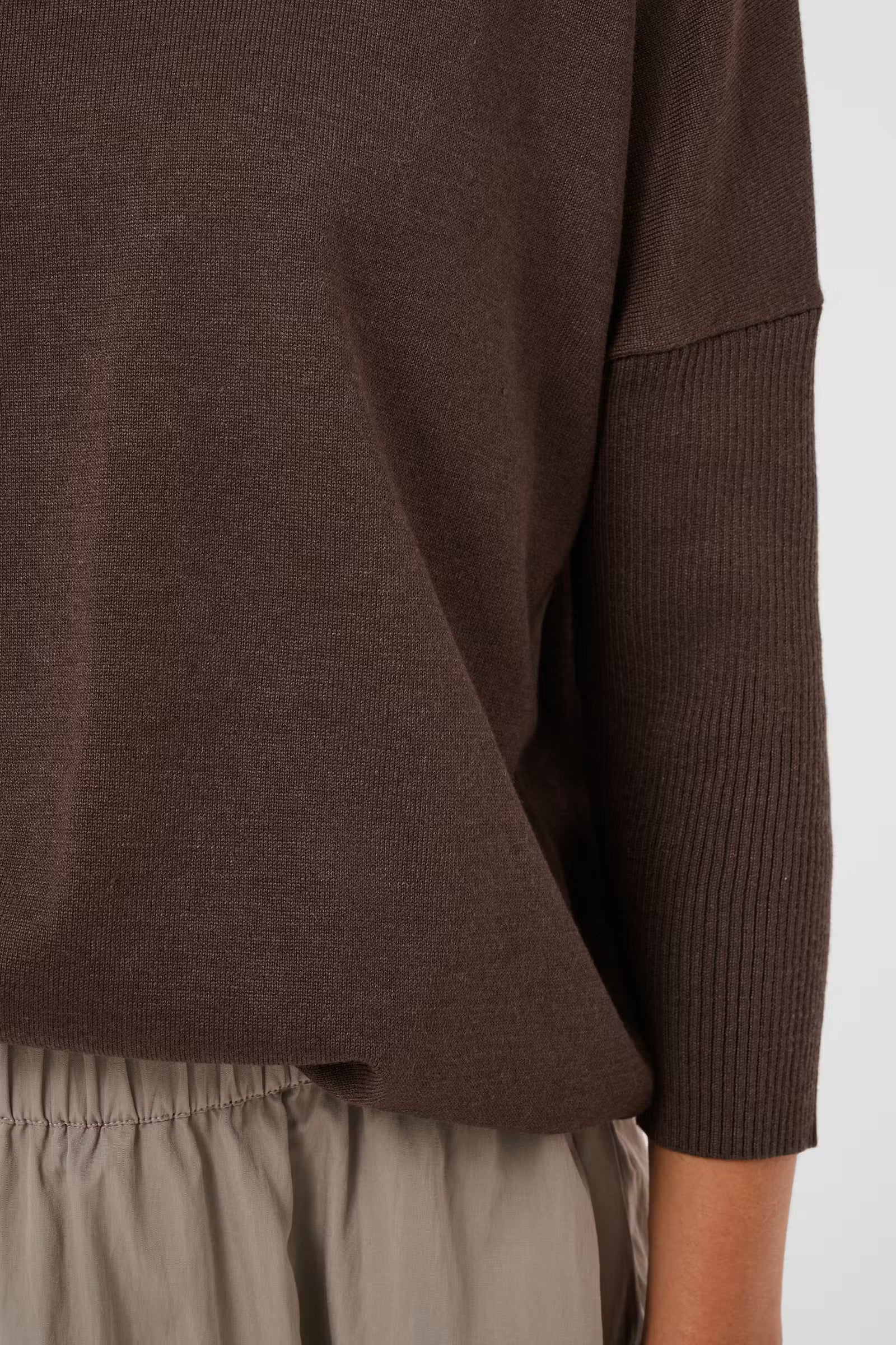 Close-up of a person wearing a dark brown knit top with a ribbed sleeve and light beige pants against a plain light background.