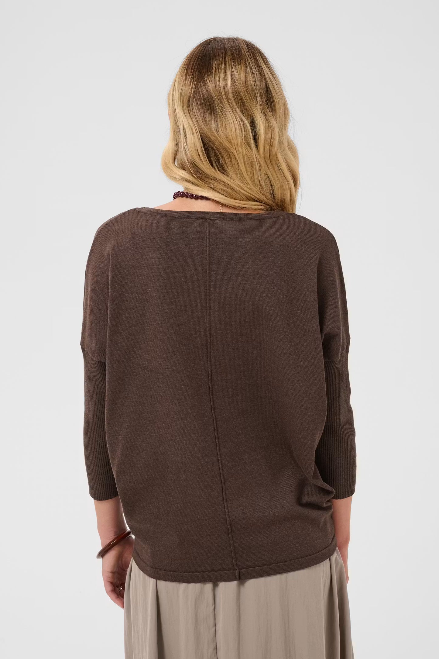 A woman viewed from the back wearing a dark brown knit top with three-quarter sleeves and a wide boat neckline, paired with light beige trousers, against a light gray studio background.