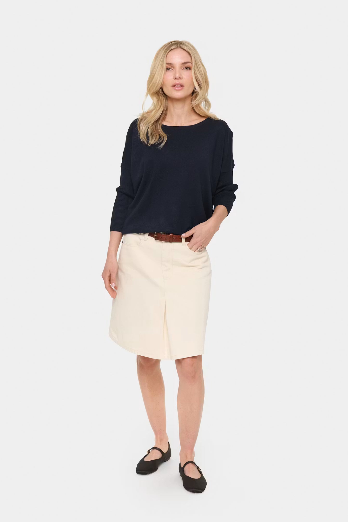 A woman modeling a dark navy blue long-sleeve top and an off-white knee-length skirt, styled with a brown belt and black flats, against a light gray studio background.