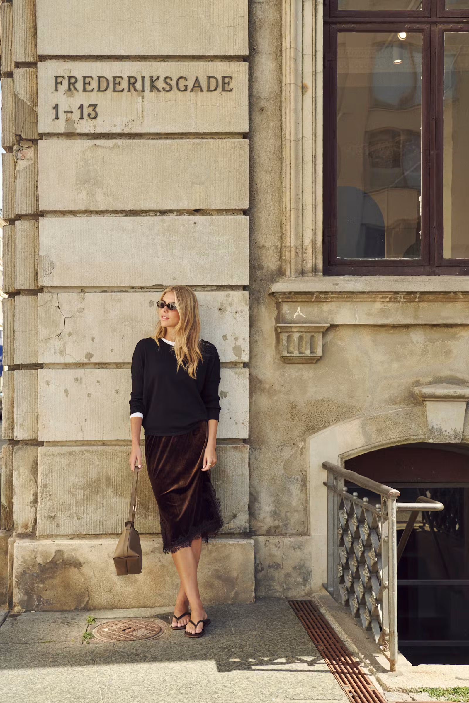 A woman stands on a city sidewalk beside a stone building facade with the text 'FREDERIKSGADE 1-13'. She is wearing a navy long-sleeve top, a dark brown velvet midi skirt, and carrying a brown shoulder bag.