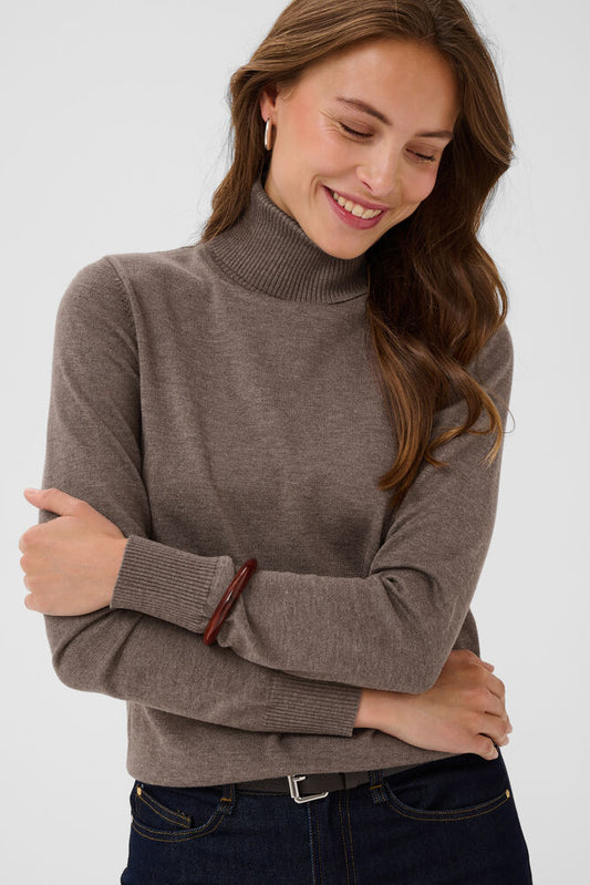A woman wearing a taupe brown ribbed turtleneck sweater and dark blue jeans, smiling and looking down against a light gray background.
