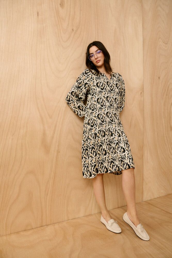 Woman wearing a patterned dress standing against a wooden paneled wall.