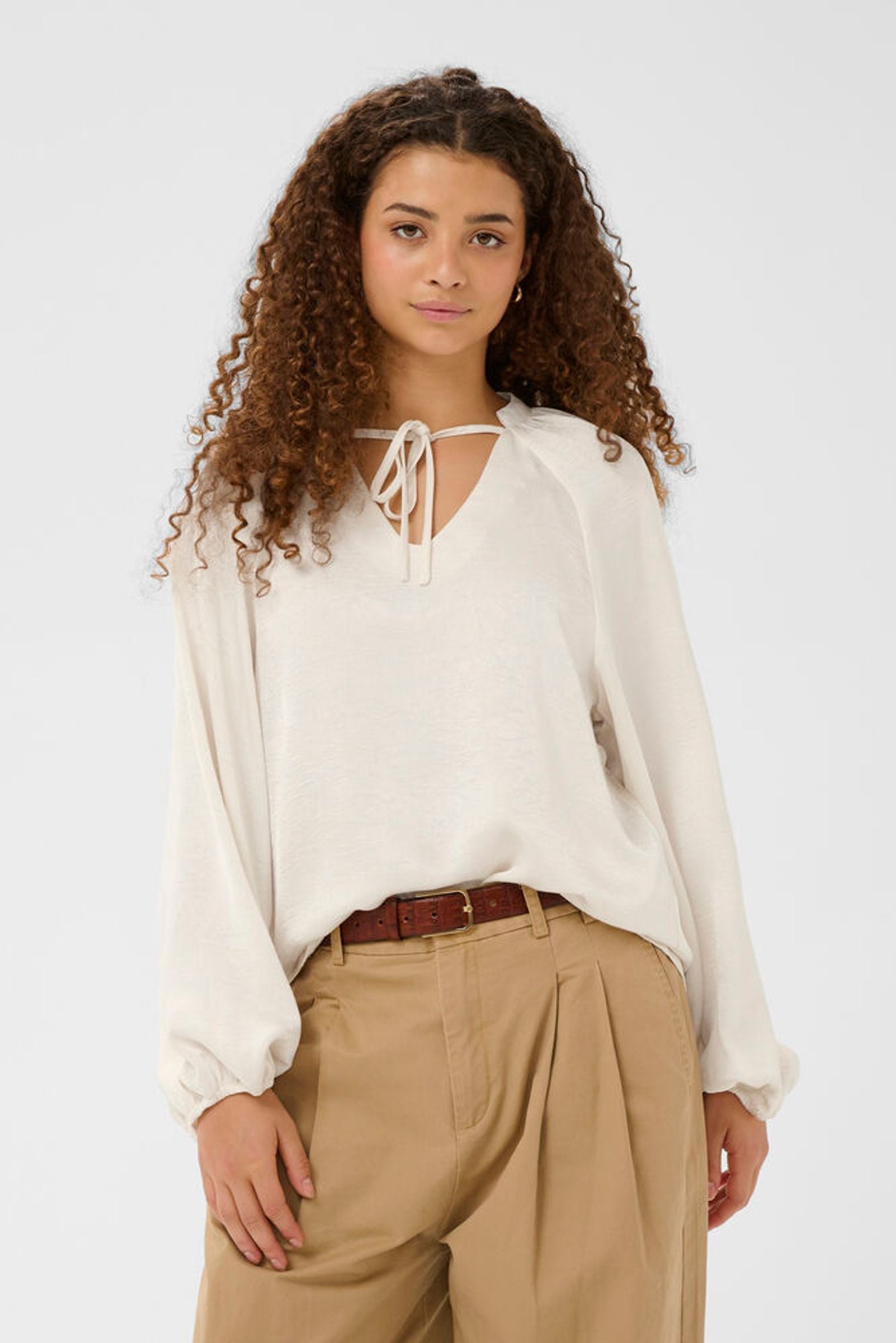 Woman wearing a white blouse with a tie neckline and beige pants on a white background