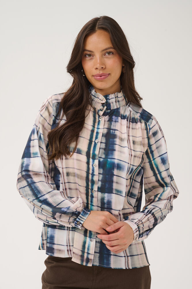 A woman modeling a long-sleeve high-neck plaid blouse with a blue, beige, and white pattern, paired with brown pants, against a plain light gray studio background.