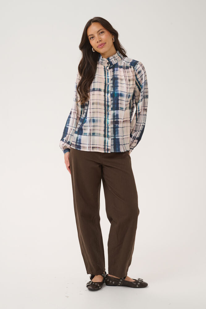 A woman modeling a long-sleeve button-up plaid shirt in blue, white, and beige, paired with brown pants and black flats, against a plain white studio background.