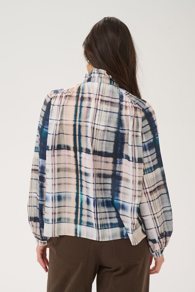 A woman shown from the back wearing a long-sleeve plaid blouse with a cream, blue, and gray pattern, paired with brown pants, against a plain light background.