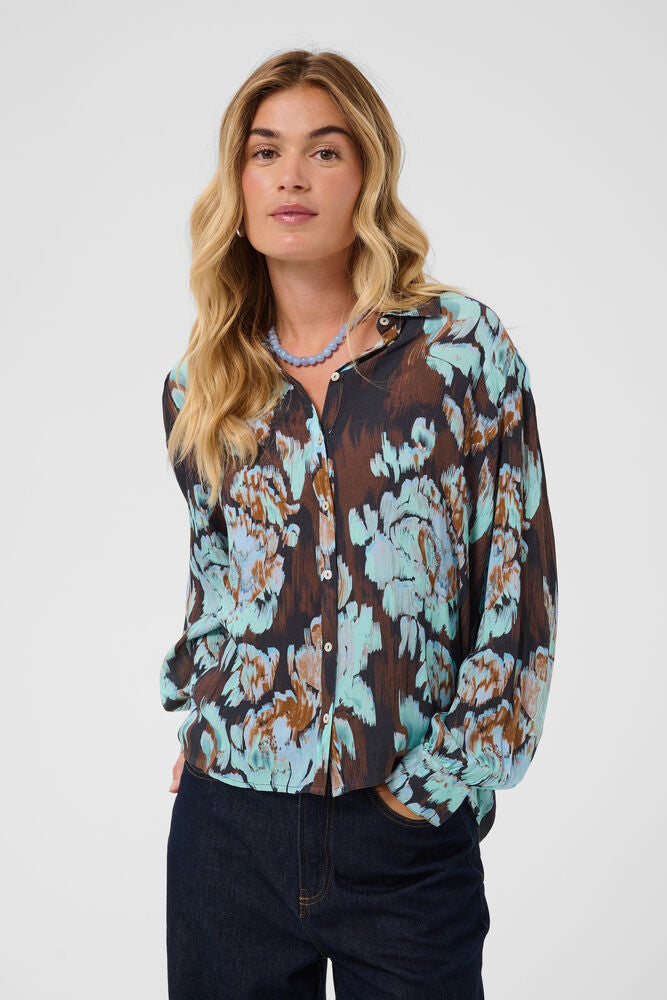 A woman wearing a long-sleeve button-up blouse with a brown, teal, and beige floral pattern, paired with dark blue jeans, against a light gray studio background.