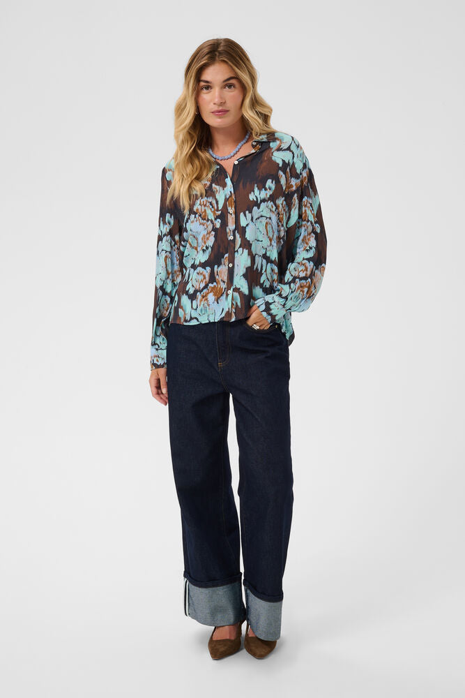 A woman modeling a sheer long-sleeve floral blouse with a brown hood, paired with dark blue jeans with rolled cuffs and brown shoes, against a light gray studio background.