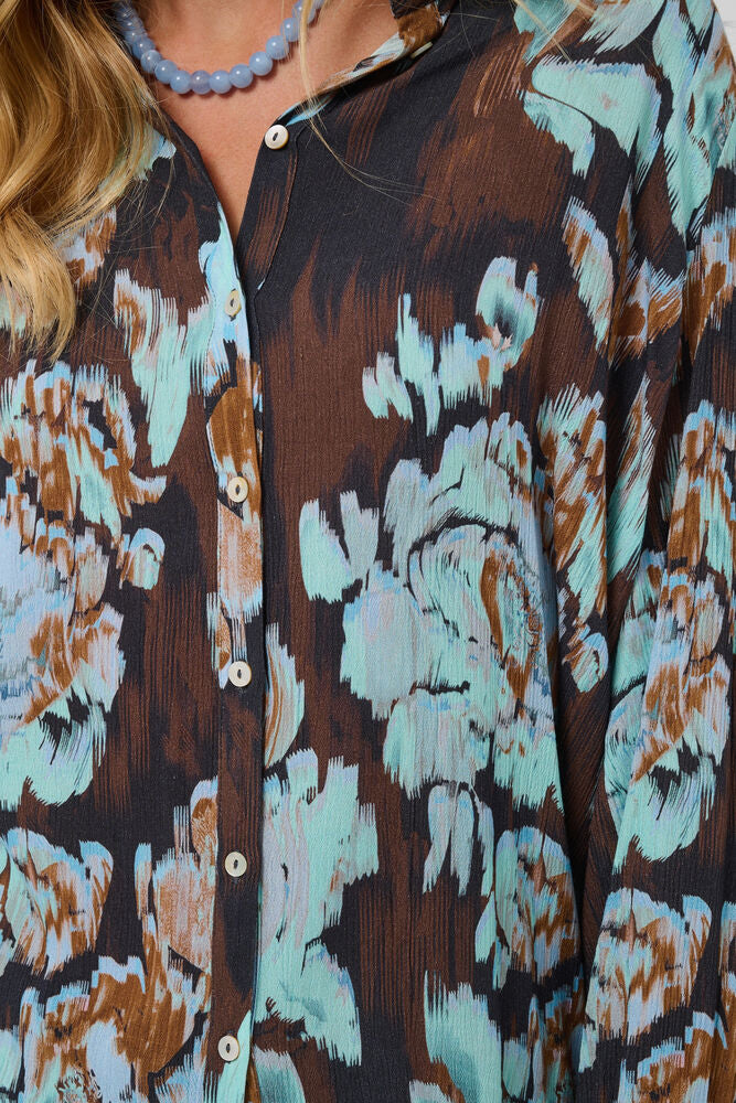 Close-up of a woman wearing a button-front blouse with a dark brown base and large abstract floral or painterly motifs in light aqua, beige, and brown.