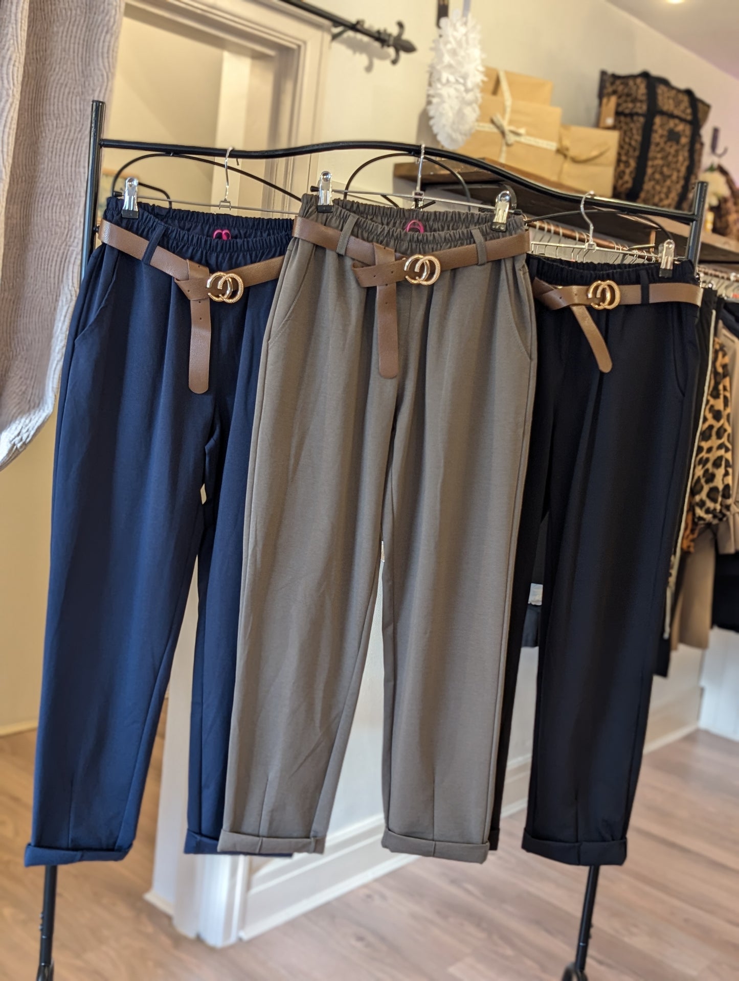 Three pairs of tailored trousers with belts displayed on hangers, in navy blue, gray, and brown colors.