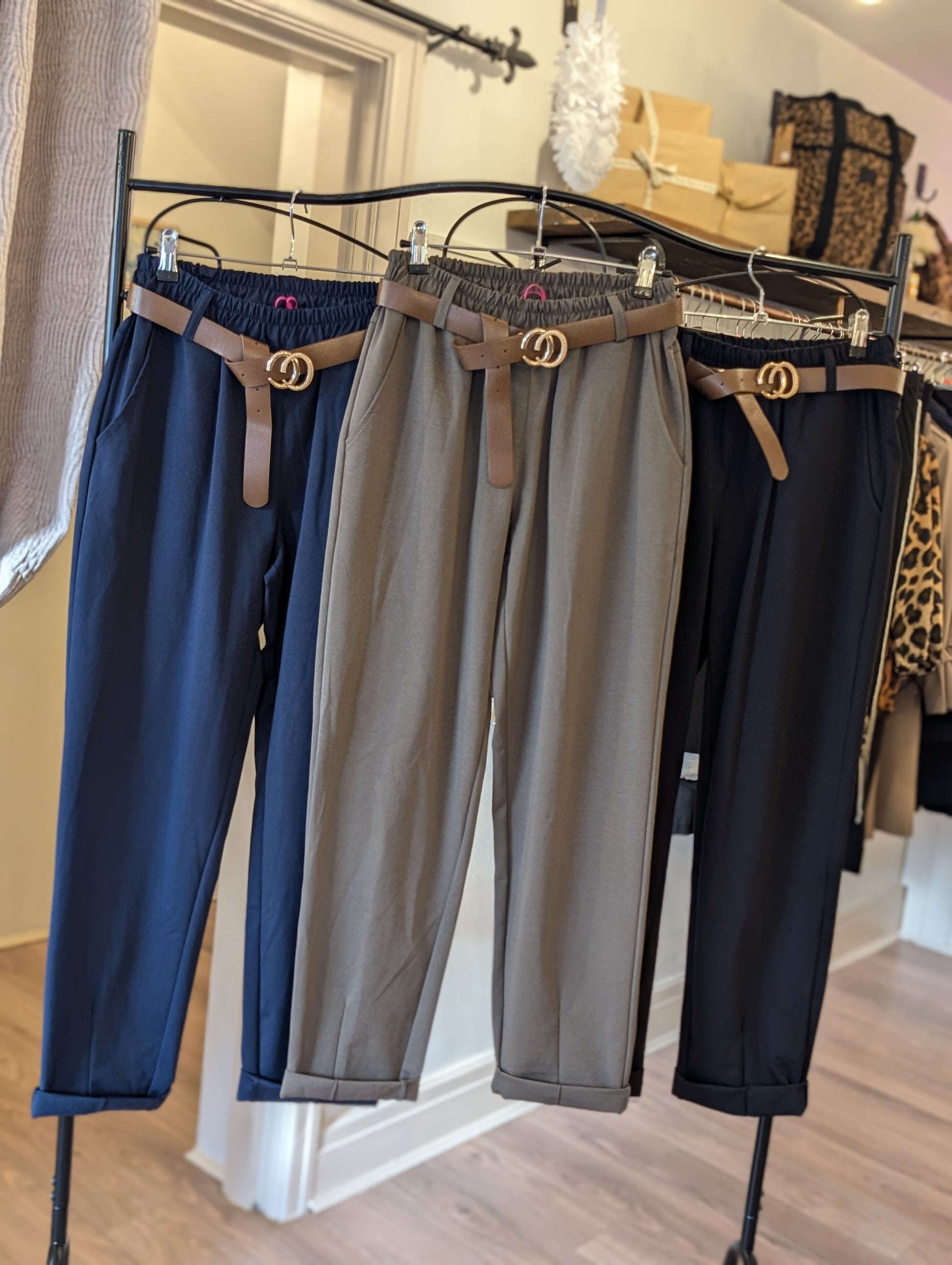 Three pairs of tailored trousers with belts displayed on hangers, in navy blue, gray, and brown colors.