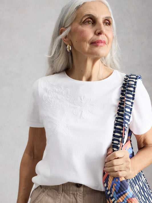 Woman with white hair wearing a white shirt holding a colorful bag against a gray background