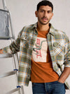 Man wearing a plaid shirt over a graphic t-shirt, standing next to a ladder.