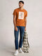 Man wearing an orange t-shirt with a graphic design, blue jeans, and white sneakers, holding a plaid shirt.