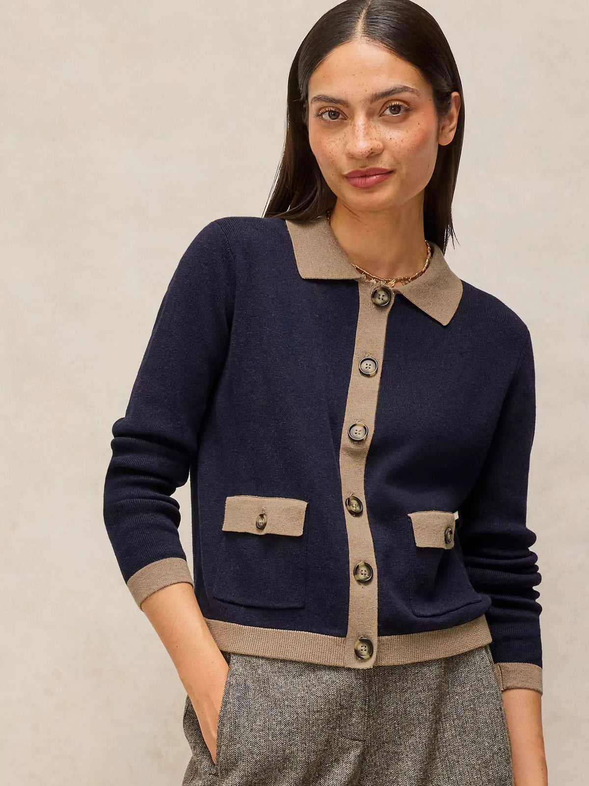 A woman wearing a navy blue knit cardigan with beige trim, featuring a pointed collar, button-front closure, and flap pockets, paired with gray textured trousers against a neutral beige background.