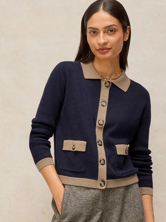 A woman wearing a navy blue knit cardigan with beige trim, featuring a pointed collar, button-front closure, and flap pockets, paired with gray textured trousers against a neutral beige background.