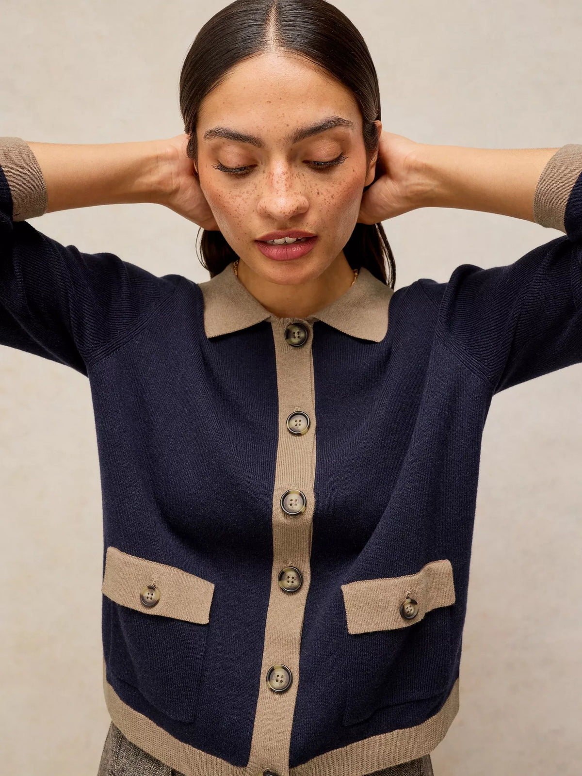 A woman wearing a navy blue knit cardigan with beige trim, a beige collar, and beige button placket, posing with her hands behind her head against a neutral beige background.