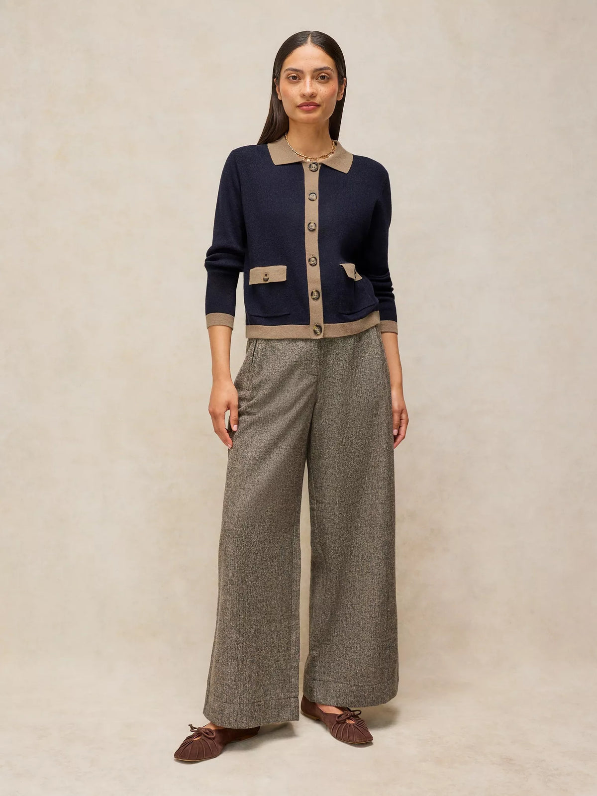 A woman stands against a warm beige studio backdrop wearing a navy cardigan with beige trim and buttons, paired with wide-leg gray trousers and brown moccasin-style shoes.