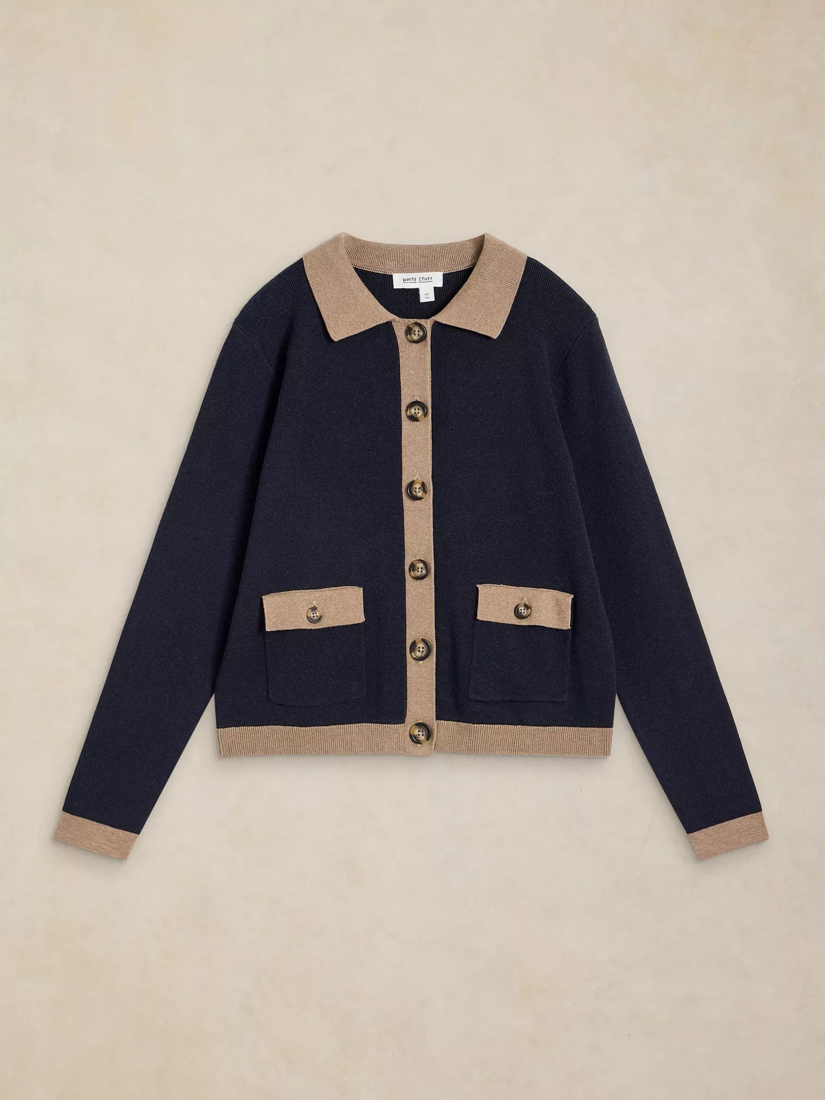 A navy blue knit cardigan with beige contrast trim on the collar, button placket, cuffs, and hem, featuring two front flap pockets with button closures, laid flat against a textured beige background.