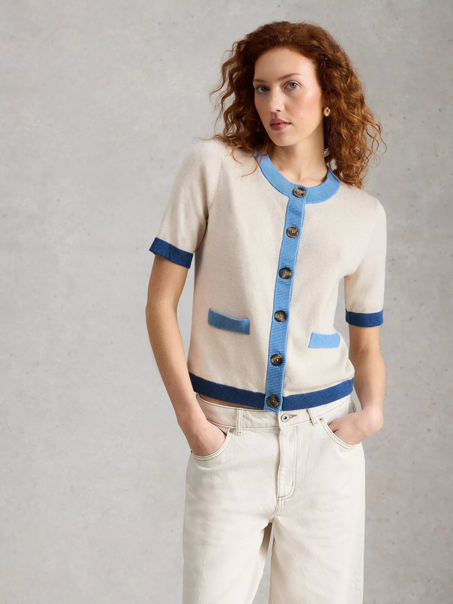 Woman wearing a beige and blue cardigan with white pants against a gray background