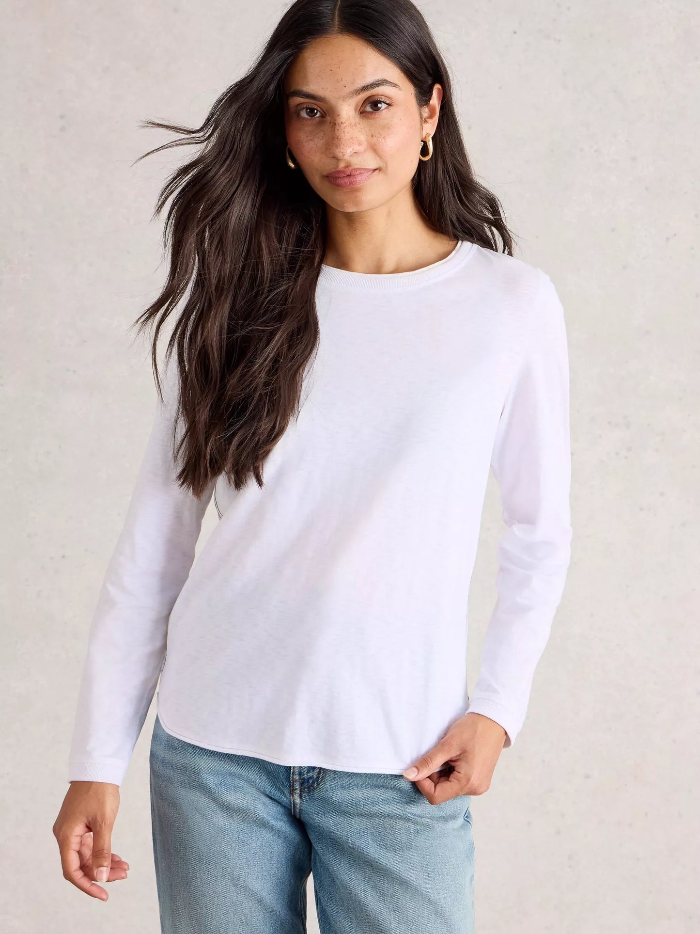 Woman wearing a white long-sleeve top and blue jeans against a neutral background