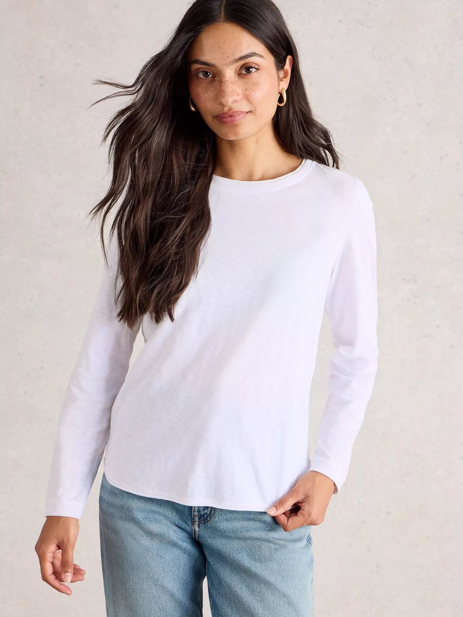 Woman wearing a white long-sleeve top and blue jeans against a neutral background