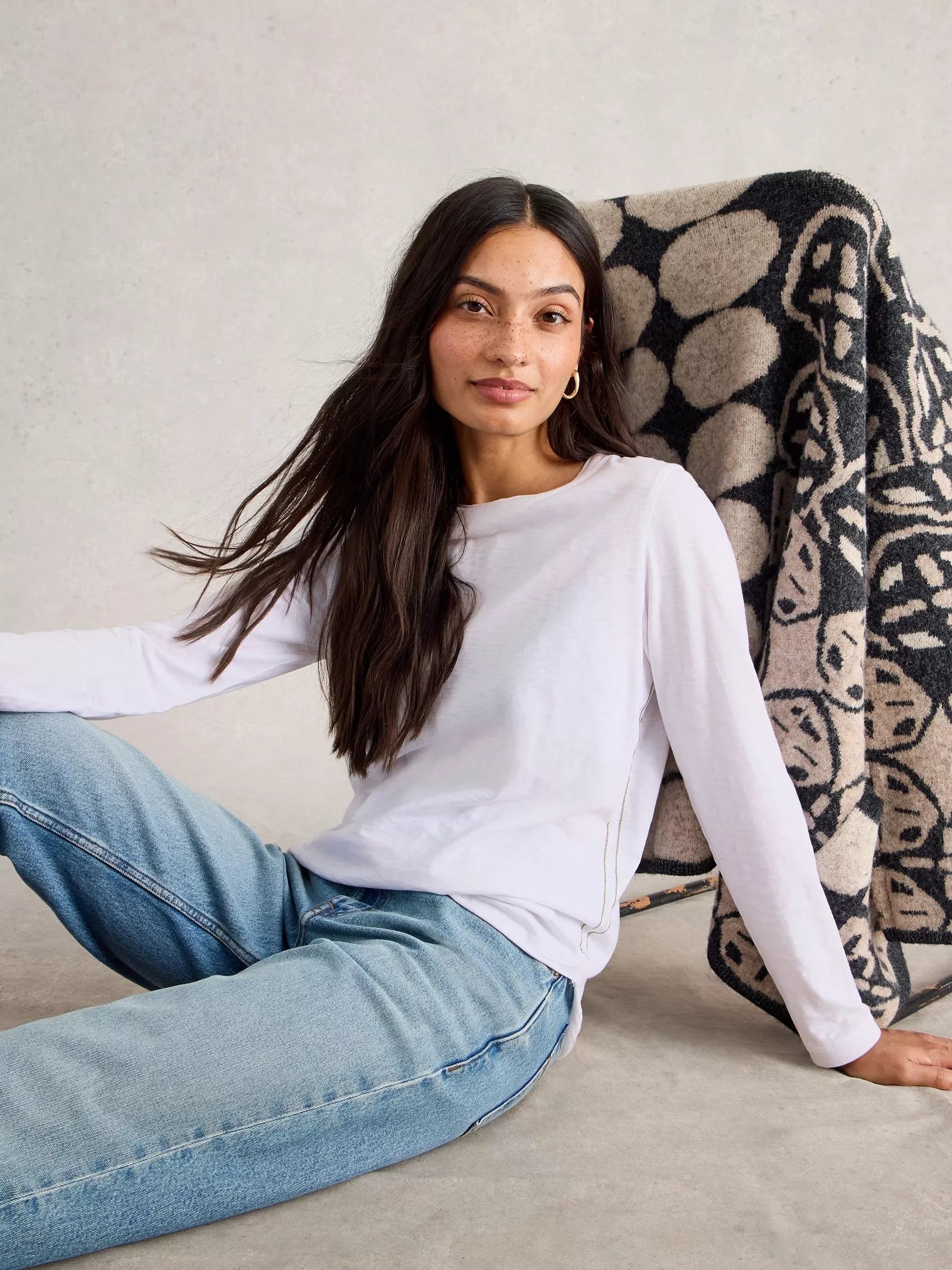Woman wearing a white long-sleeve top and blue jeans sitting on a couch with a patterned blanket.