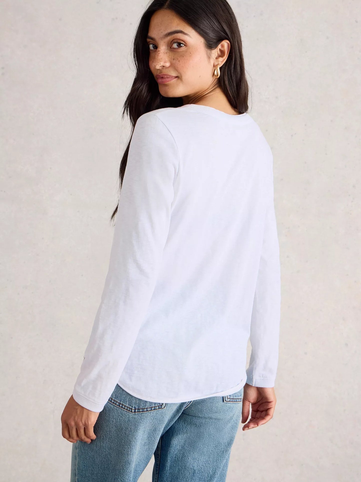Woman wearing a white long-sleeve shirt and blue jeans against a plain background