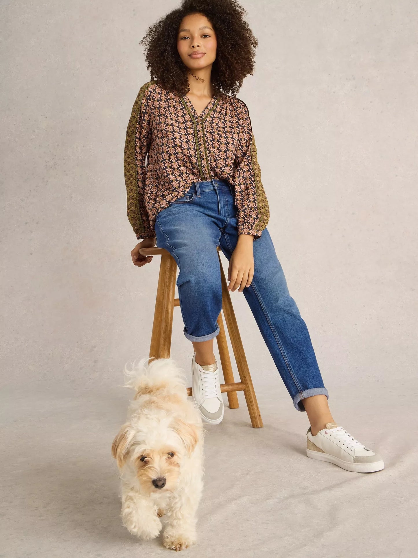 Woman sitting on a stool with a dog at her feet, wearing a patterned top and jeans.