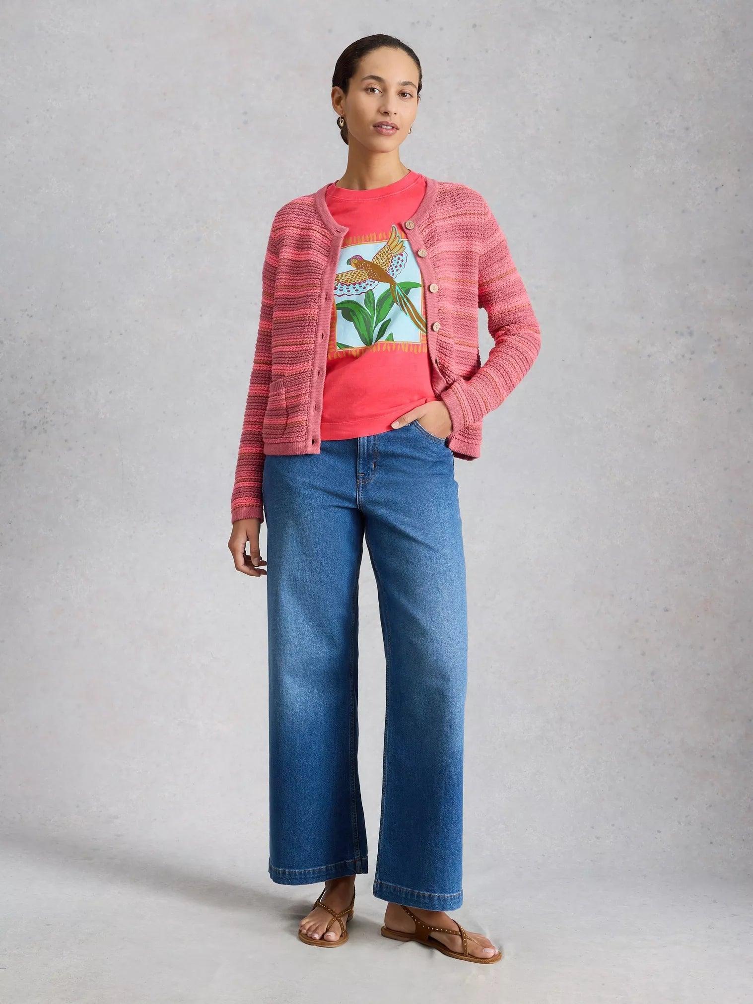 Woman wearing a pink cardigan, red shirt with graphic design, and blue jeans on a gray background