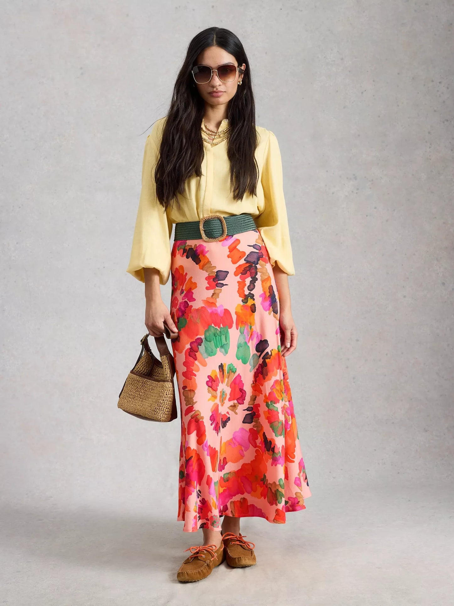 Woman wearing a yellow blouse, colorful floral skirt, and brown shoes on a gray background