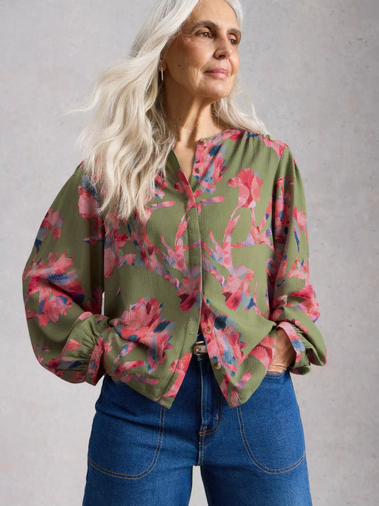 Woman wearing a floral blouse with green base and pink flowers, paired with blue jeans against a plain background.