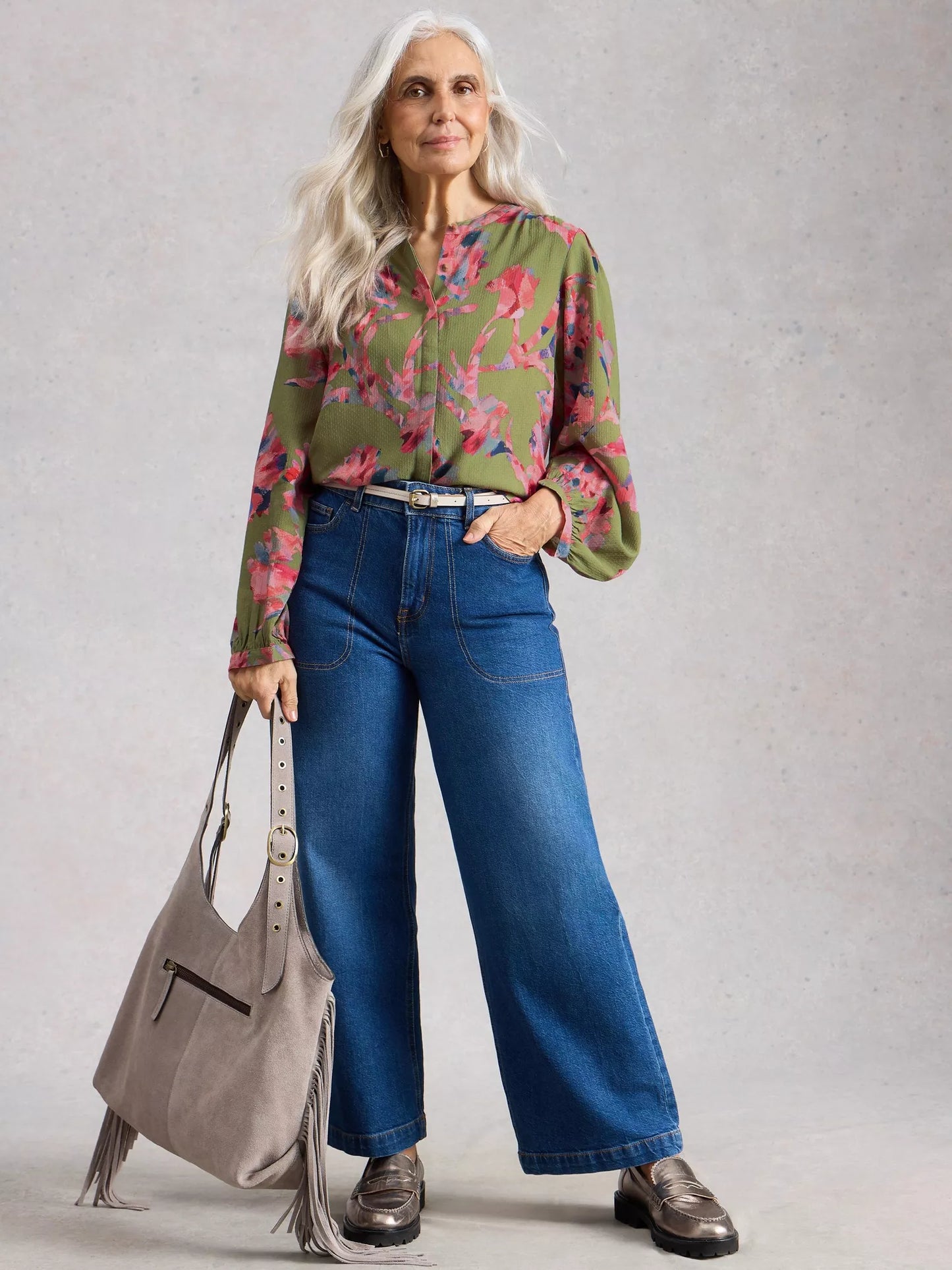 Woman wearing a floral blouse and blue jeans, holding a gray handbag against a plain background