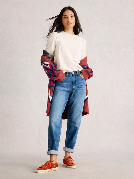 Woman wearing a colorful cardigan, white shirt, blue jeans, and red shoes on a plain background