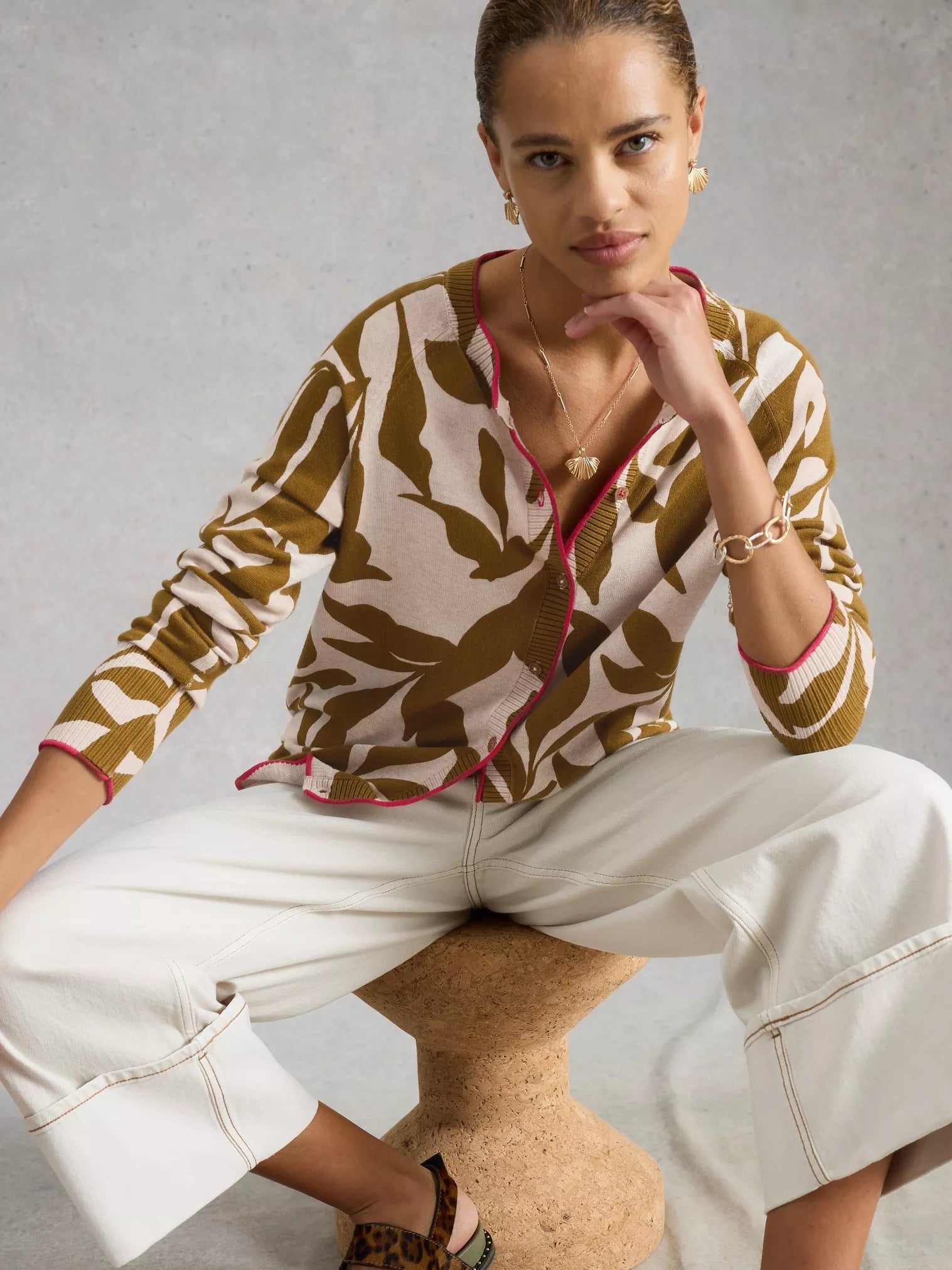 Woman wearing a patterned top and white pants sitting on a chair against a neutral background