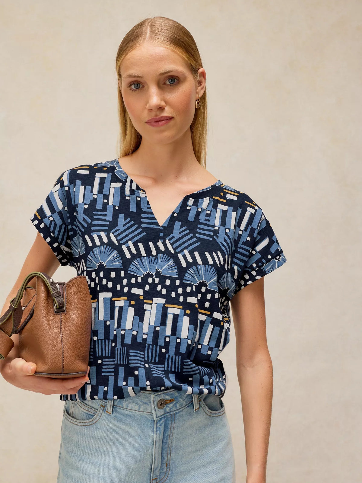 Woman wearing a blue patterned top and holding a brown leather bag against a beige background