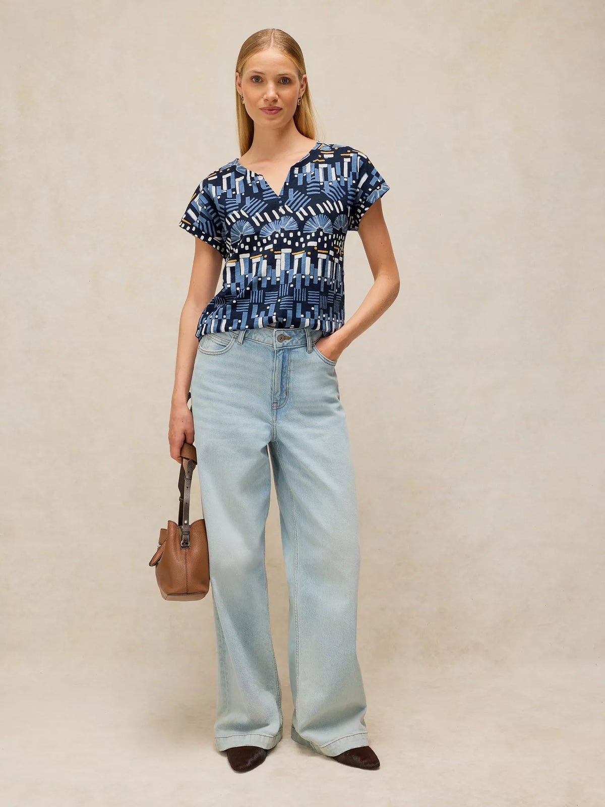 A woman stands against a warm beige textured backdrop wearing a blue and white geometric patterned short-sleeve top, light-wash wide-leg jeans, and dark brown pointed shoes, while holding a small brown handbag.