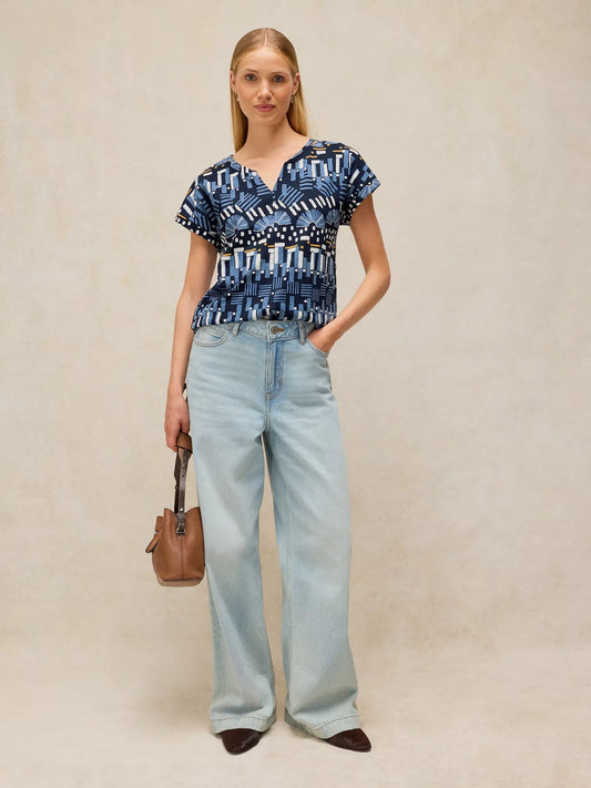 A woman stands against a warm beige textured backdrop wearing a blue and white geometric patterned short-sleeve top, light-wash wide-leg jeans, and dark brown pointed shoes, while holding a small brown handbag.
