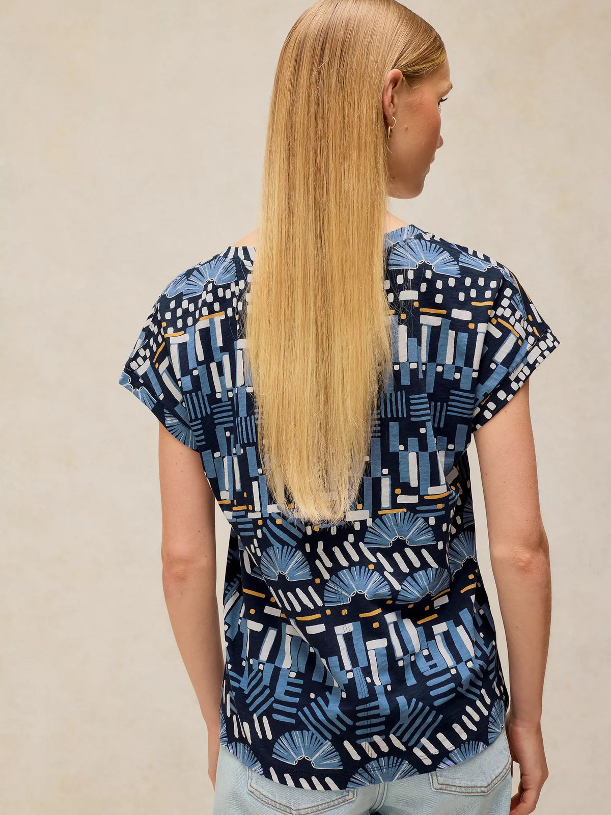 A woman with long blonde hair is shown from the back, wearing a short-sleeve top with a navy blue, white, and light blue geometric and fan-like pattern, paired with light blue jeans against a neutral beige background.