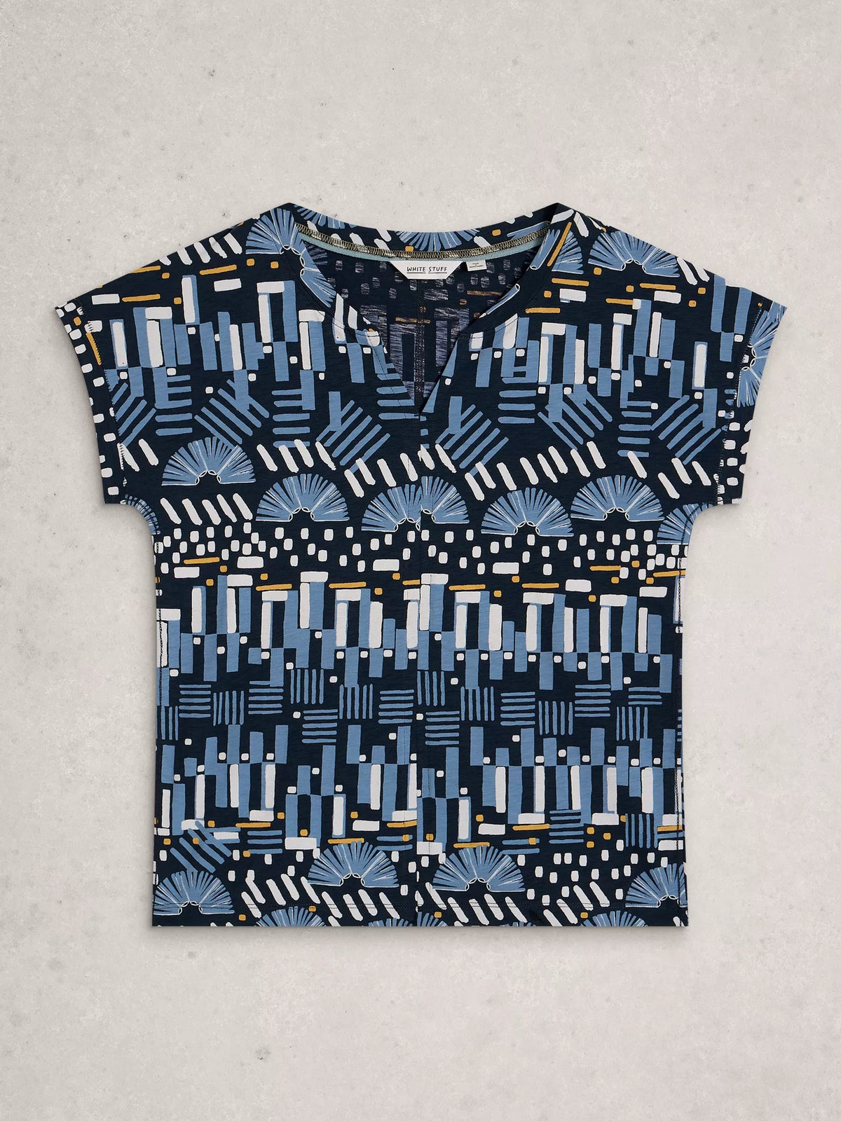A short-sleeve crewneck top with a geometric and fan-like abstract pattern in navy, light blue, white, and small yellow accents, laid flat on a light gray textured background.