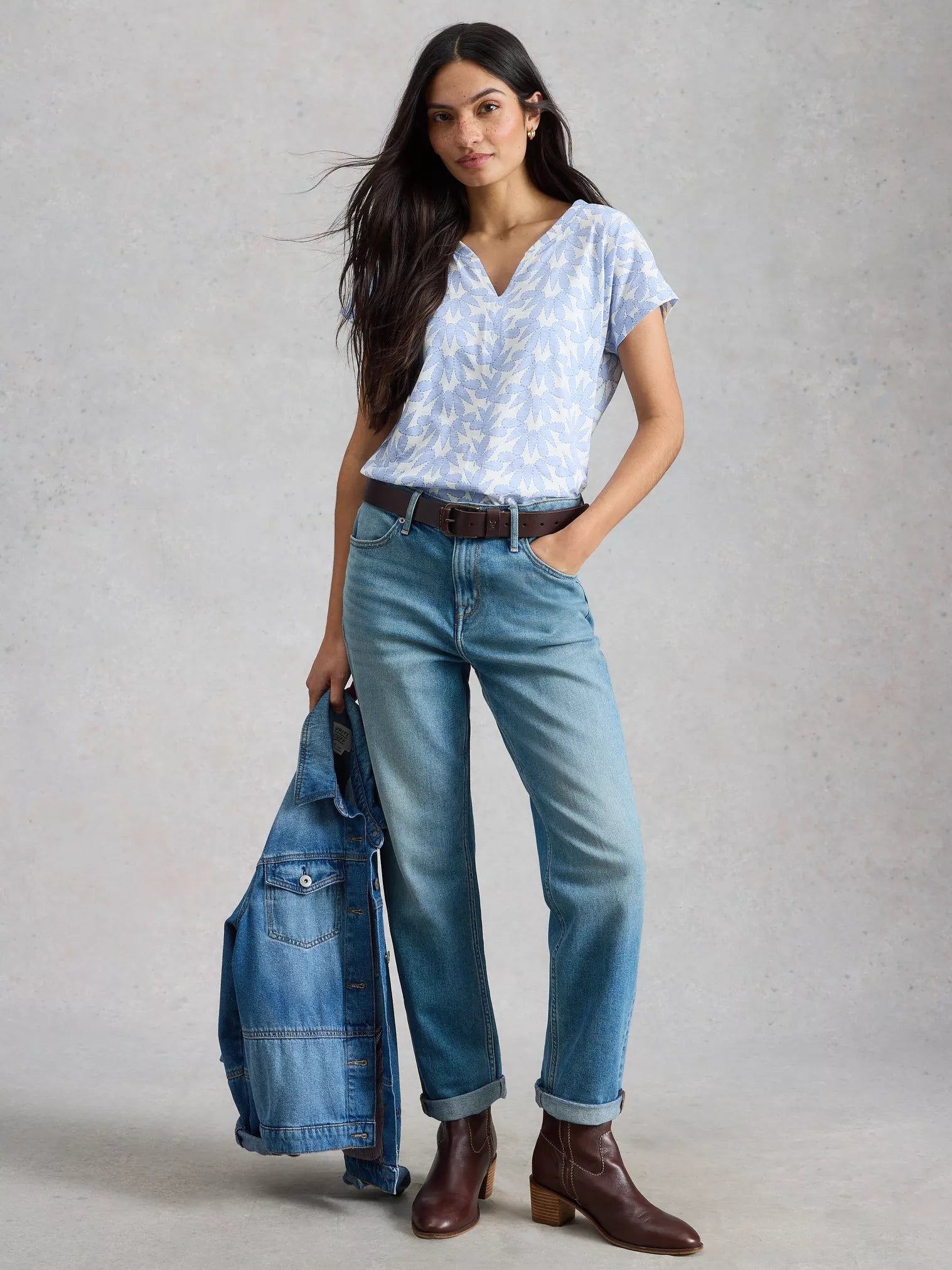 Woman wearing a light blue floral blouse, jeans, and brown boots holding a denim jacket.